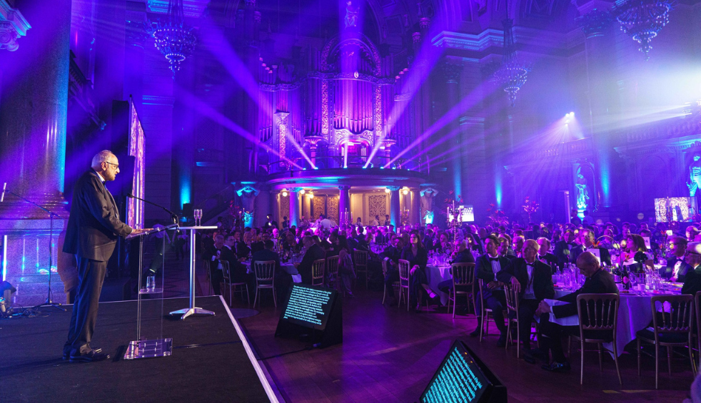 Person standing at a lectern on a stage, speaking to a large audience seated at round tables in a grand hall lit with purple and blue stage lighting. The venue features tall columns, chandeliers, and a large pipe organ at the far end, with spotlights and illuminated screens highlighting the formal event setting.