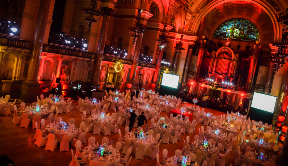 Large, ornate hall set up for a formal event, with dozens of round tables covered in white linens and glowing centerpieces. The room is lit with dramatic red lighting, featuring tall columns, chandeliers, and a grand pipe organ at the far end next to two large projection screens.