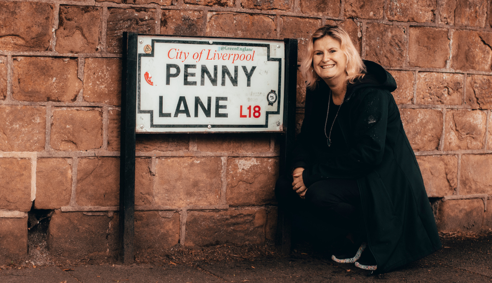A person dressed in a long black coat and dark outfit crouches beside a mounted street sign reading “City of Liverpool – Penny Lane L18,” which is fixed to a weathered red brick wall. The ground appears damp, and the scene is captured in warm, muted tones, with the person positioned to the right of the sign and facing the camera.