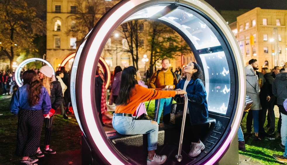 Visitors gather around illuminated circular light installations in a city square at night. One large glowing pod-like structure in the foreground contains seating and interactive light displays, while additional illuminated rings can be seen throughout the crowd. Historic buildings and trees form the backdrop, with colourful lighting creating a vibrant festival atmosphere.