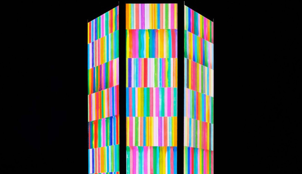 Three tall rectangular panels illuminated with vibrant vertical stripes in rainbow colours stand against a dark night sky. The colourful light installation features bands of pink, blue, green, yellow, orange and purple, creating a bright geometric display that contrasts sharply with the surrounding darkness.