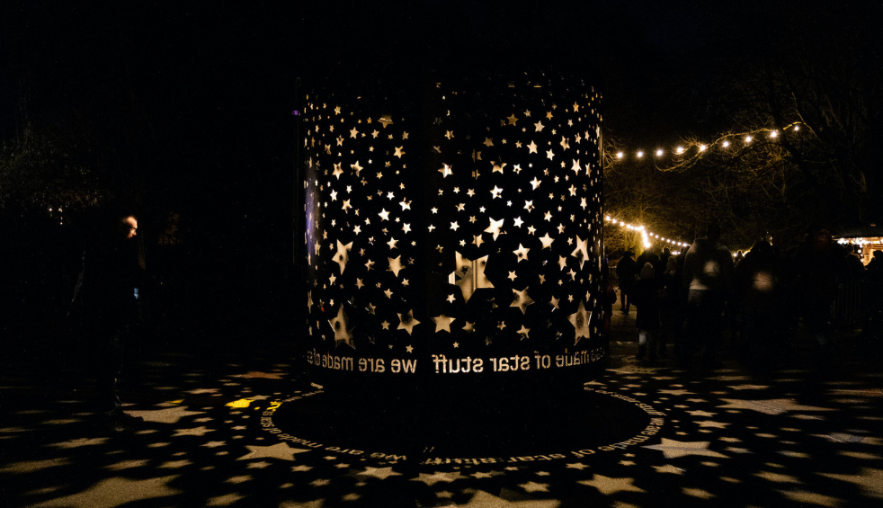 A large cylindrical lantern perforated with star-shaped cut-outs glows in a dark outdoor setting, casting illuminated star patterns and text onto the ground. Strings of warm white lights hang overhead, while visitors walk along a path in the background, creating a festive nighttime atmosphere.