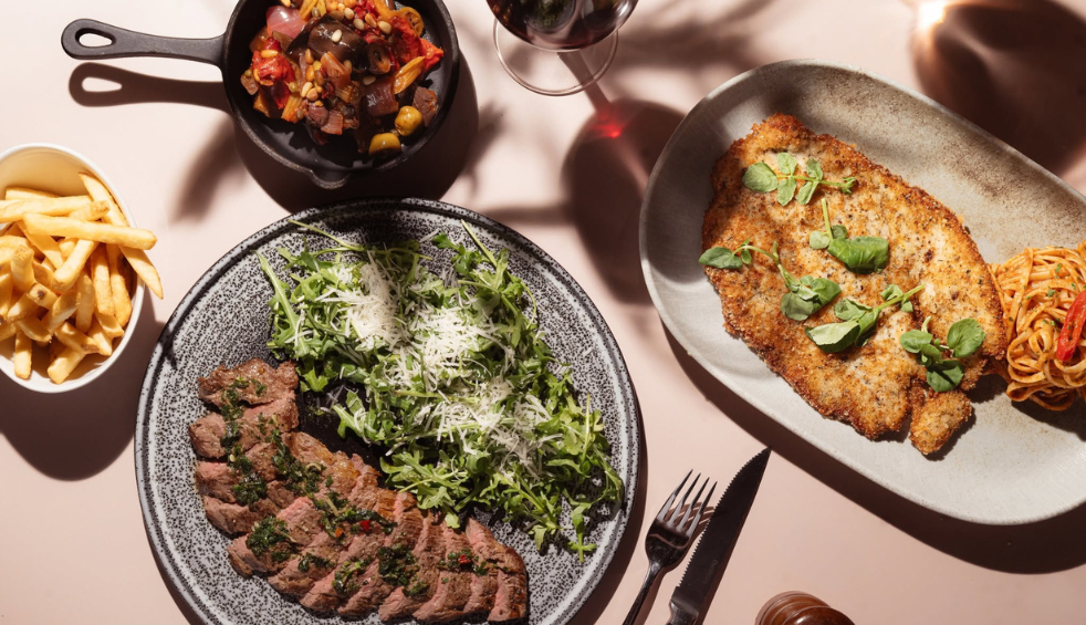 Overhead view of a restaurant table featuring a selection of dishes, including sliced steak served with a leafy salad topped with grated cheese, a breaded chicken fillet garnished with fresh herbs and spaghetti, a bowl of fries, and a cast-iron skillet filled with roasted vegetables. A glass of red wine and cutlery complete the dining scene.