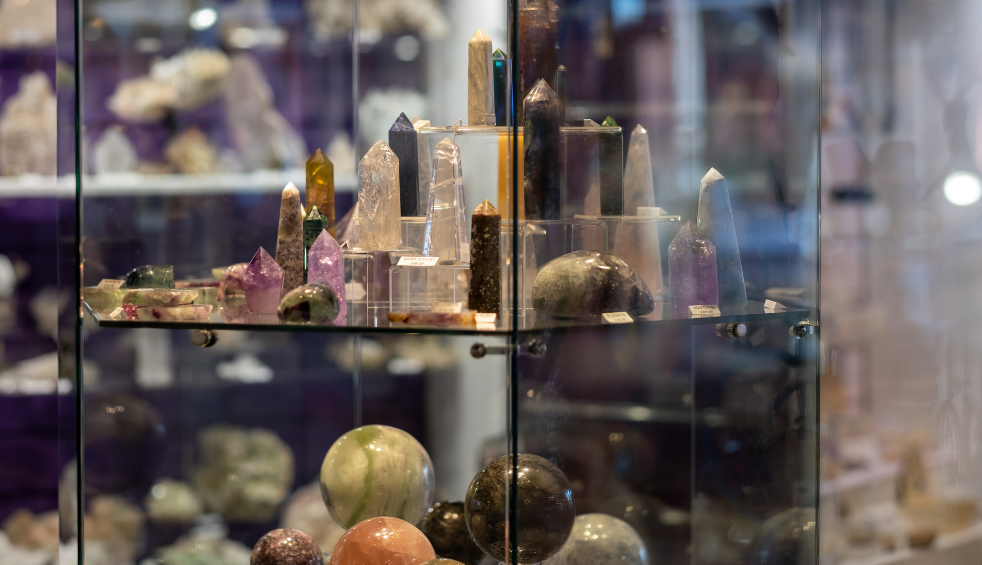 A glass display cabinet filled with polished crystals, mineral specimens and gemstone spheres. Various crystal points in clear, purple, green and dark tones are arranged on glass shelves, while larger stone spheres sit below. The softly lit interior and blurred background create a museum or crystal shop setting.