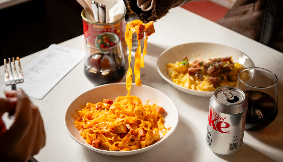 A restaurant table featuring bowls of pasta and other dishes, with a fork lifting strands of creamy tagliatelle from a plate in the foreground. A can of Diet Coke, glasses of soft drink, and additional meals including pasta and meat dishes are arranged around the table, creating a casual dining scene.