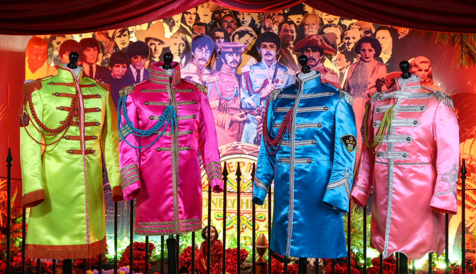 Four brightly coloured satin military-style band jackets displayed on mannequins, featuring lime green, pink, blue and pale pink designs with decorative braiding and shoulder cords. The costumes are set against a vibrant backdrop of illustrated figures and album artwork associated with Sgt. Pepper’s Lonely Hearts Club Band, under red draped fabric and theatrical lighting.
