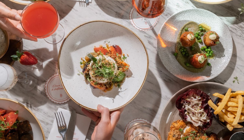Overhead view of a marble-topped restaurant table set with a selection of contemporary dishes and drinks. A central plate features burrata with tomatoes and herbs, surrounded by croquettes served with peas, fries, coleslaw, and cocktails. Hands reach in to hold a plate and a pink-coloured drink, creating a shared dining atmosphere.