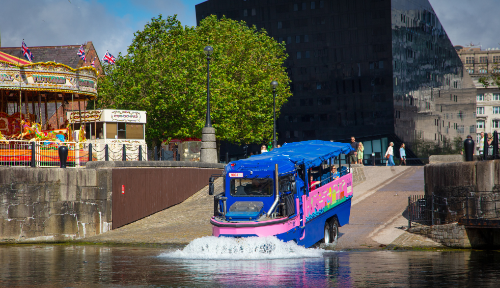 A brightly coloured blue and pink amphibious tour vehicle enters the water from a cobbled slipway at Liverpool’s waterfront. Trees, a vintage carousel and modern buildings form the backdrop as the vehicle creates a splash entering the dock.