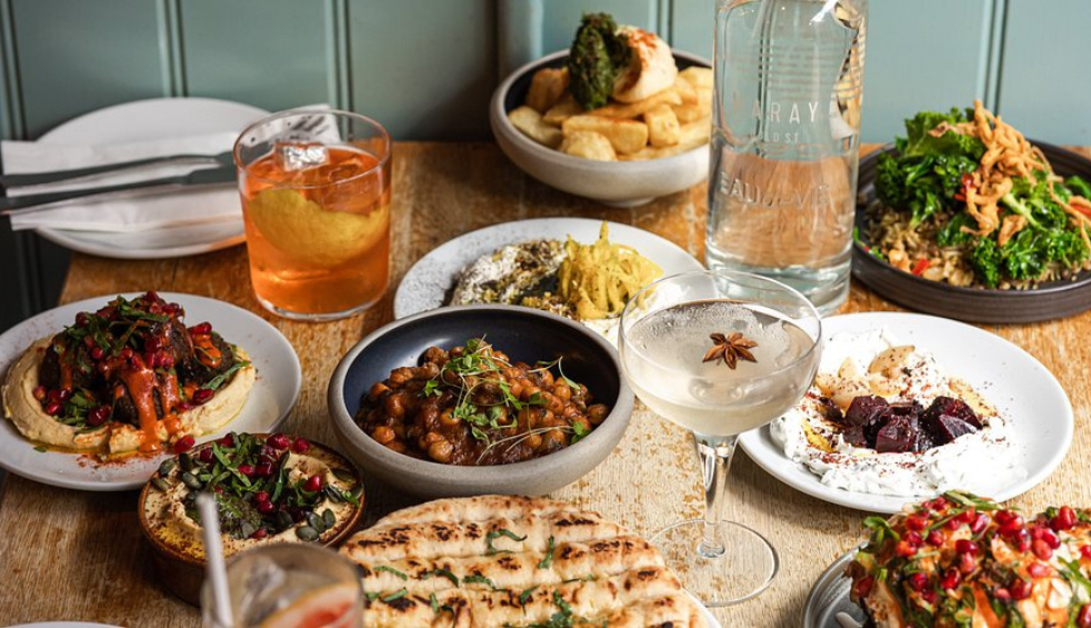 A table filled with colourful sharing plates, flatbreads, salads, dips and cocktails in a restaurant setting. The dishes are arranged for communal dining on a light wooden table.