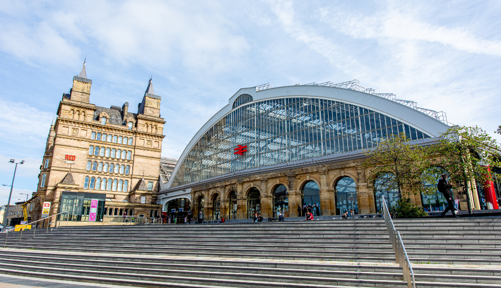 Outside of Liverpool Lime Street Station which is an arched shaped building with glass windows at the top and brick at the bottom.