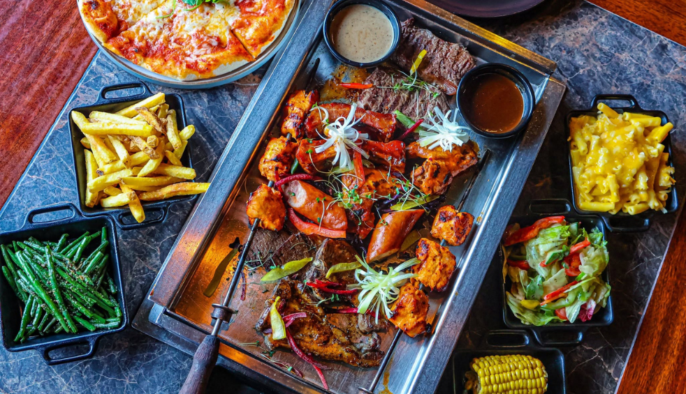 A table full of plates of food including grilled meats, pizza, vegetables, chips and salad.