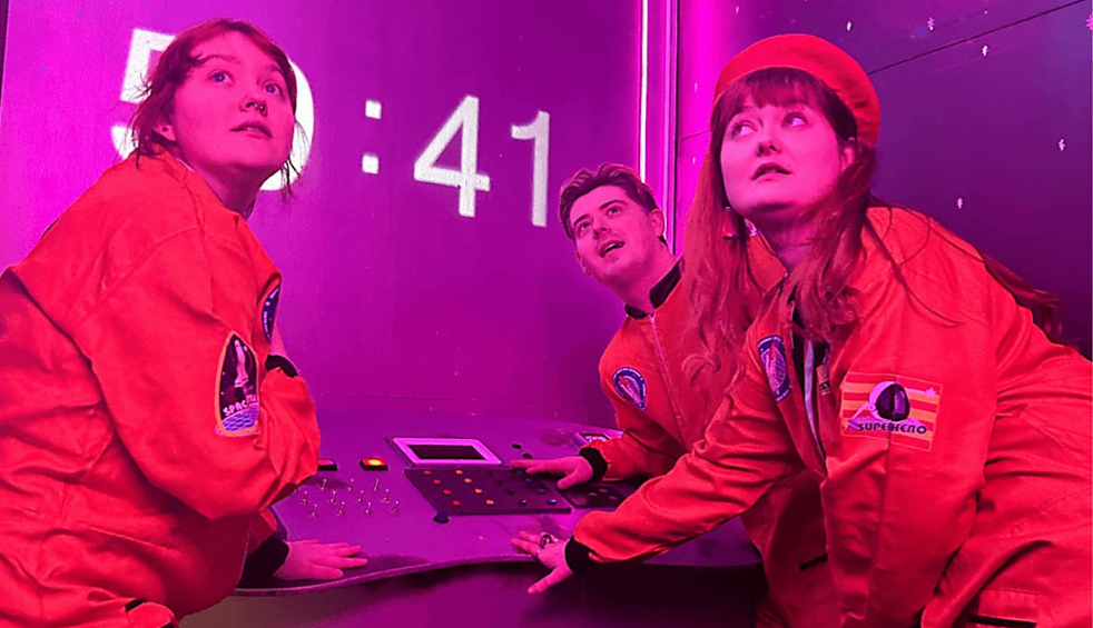 Three people wearing bright orange astronaut-style suits stand around a control panel inside a neon‑lit room. Digital numbers reading “:41” glow on the wall behind them, while purple and pink lighting creates a futuristic, immersive setting.