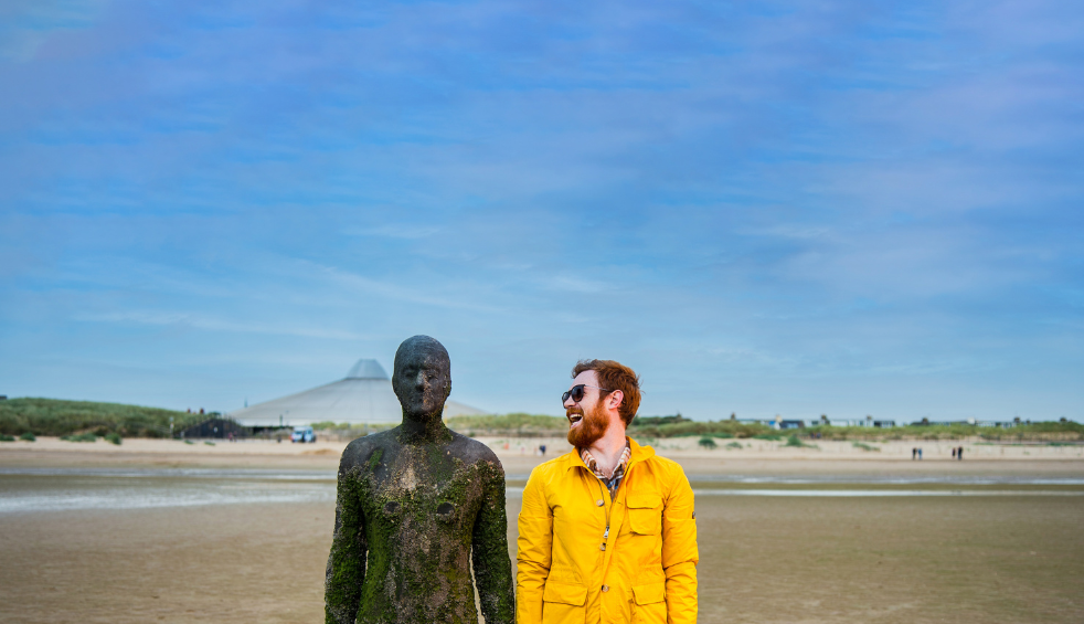 A person wearing a yellow coat stood next to an iron man statue on Crosby Beach.