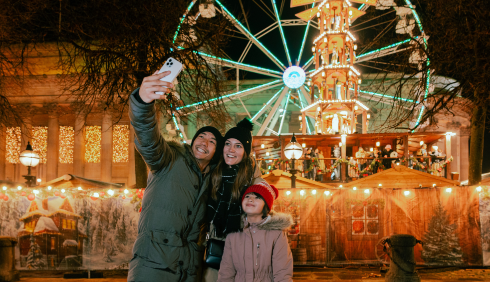 A family of three, two adults and a child taking a selfie in front of the Christmas markets in liverpool at St George's Hall.