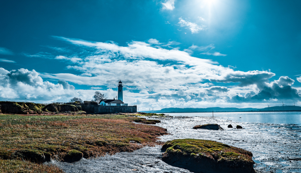 A river with a grass path that has a light house at the end of it with a blue sky.