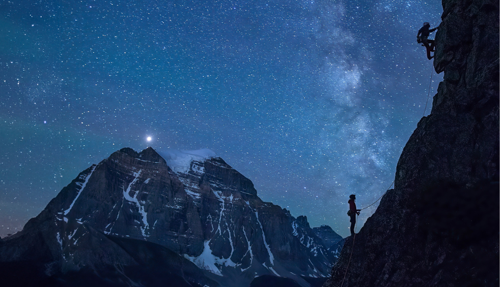 An image showing a starry night with two people climbing a mountain.