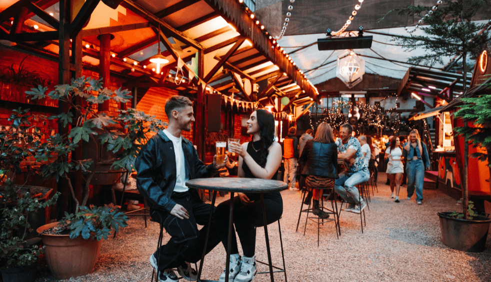 Inside a beer garden venue with string lights and two people sitting on a table with drinks.