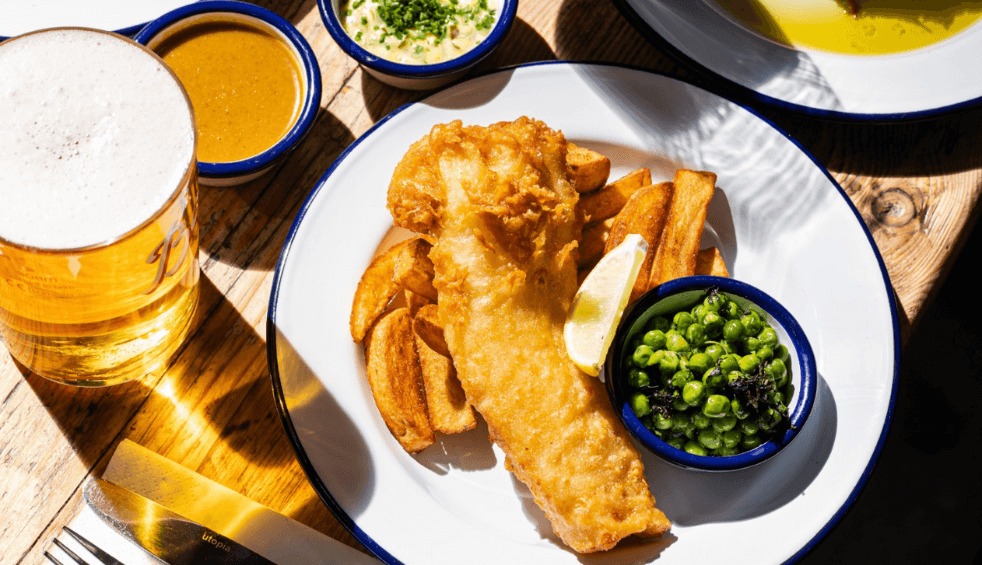 Fresh fish and chips with peas on a plate in the sunshine. There's also a pint of larger and some ramakins of sauce.
