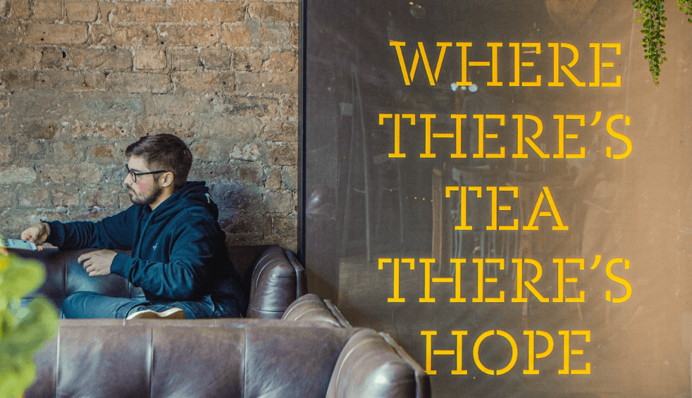 A person sitting on a couch with a cup of tea with a large sign next to them that says Where There's Tea There's Hope