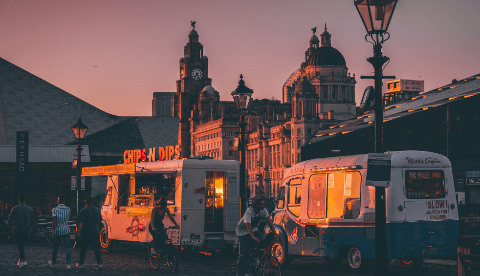 A sunset shot cross section with two retro food vans in the front with the view of the Royal Liver Building, Cunard and Port of Liverpool building in the background.