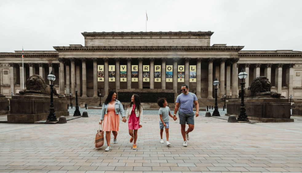 A family of four walking with their back to St George's Hall, there's banners on the hall too with a lion monument on the right and left of the family.