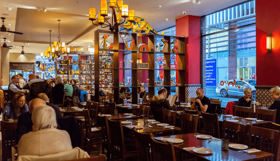 Inside a large restaurant with red walls, big windows and golden light lanterns. There's wooden tables and shelves packed with traditional Spanish items.