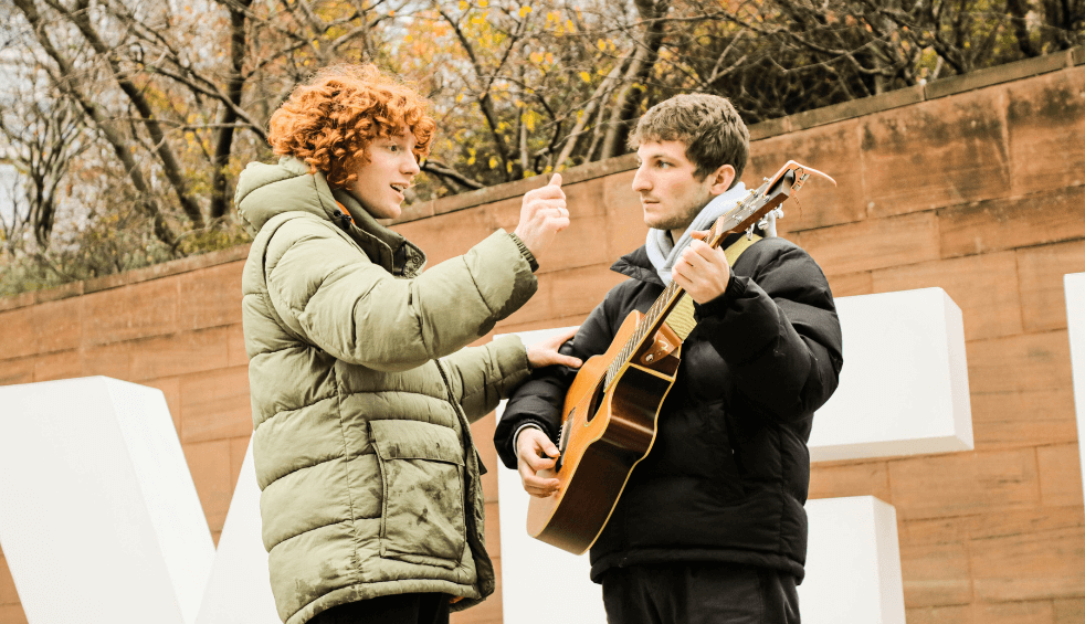 two people outside one singing and one playing an acoustic guitar