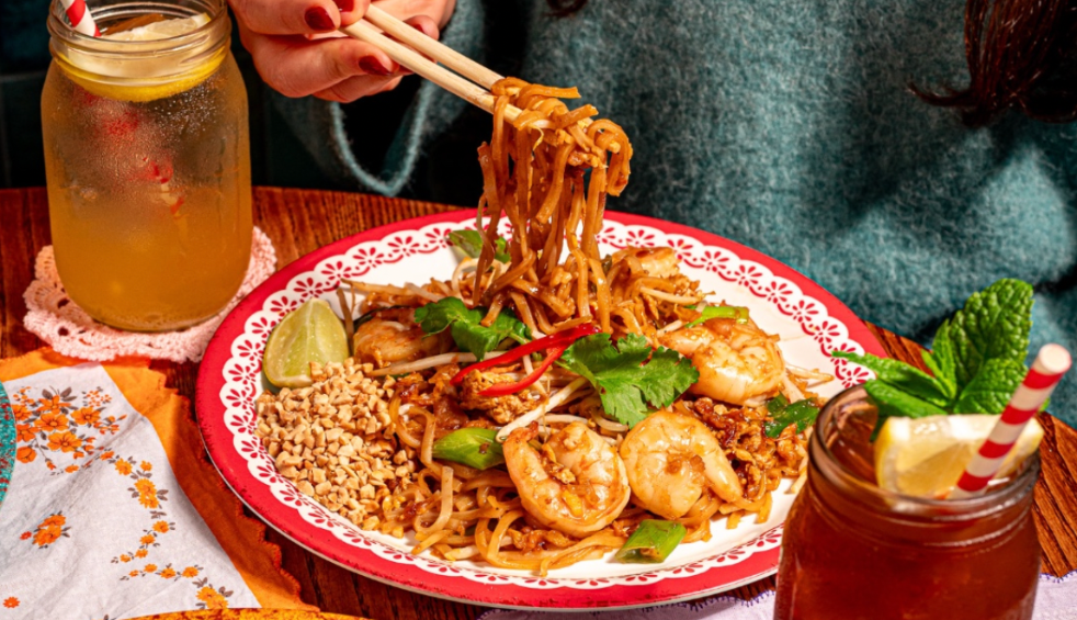 A plate with noodles and prawns on. Someone is pulling noodles off the plate with chopsticks.