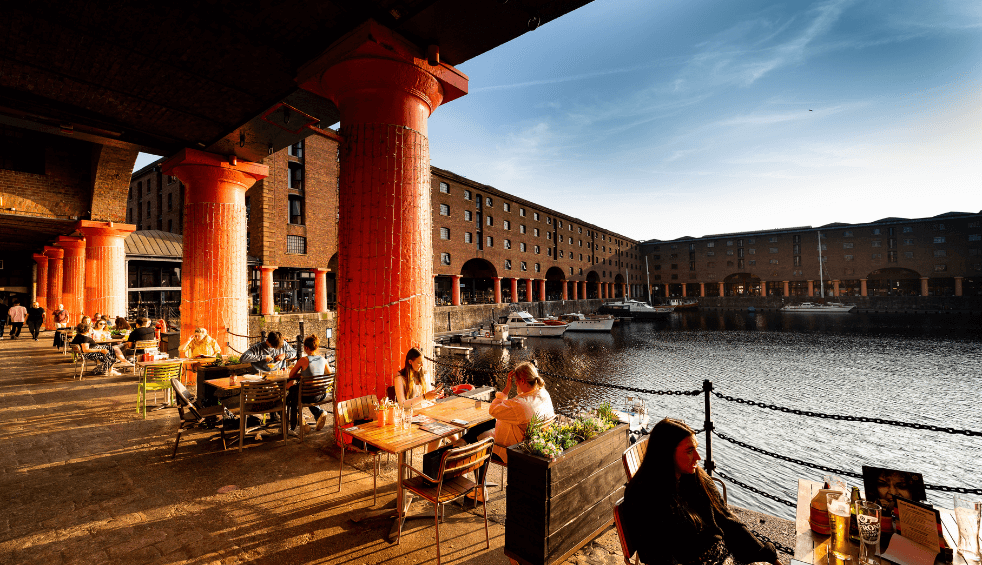 A sunny day on the Royal Albert Dock - People are sitting at tables by large, red pillars overlooking the dock,