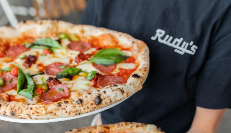A pizza with cheese, pepperoni and basil on it being held by someone wearing a black top that says Rudy's.