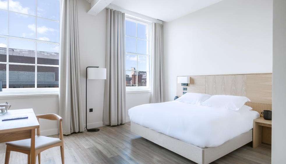 Inside a hotel room with a large double bed in the middle of the room. The bedding is white. There are two large windows on the wall.