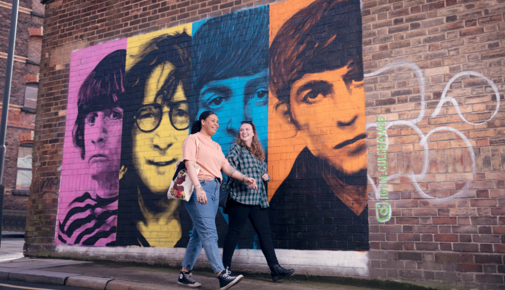 Two girls walk past a pink, yellow, blue and orange street art portrait of the Beatles.