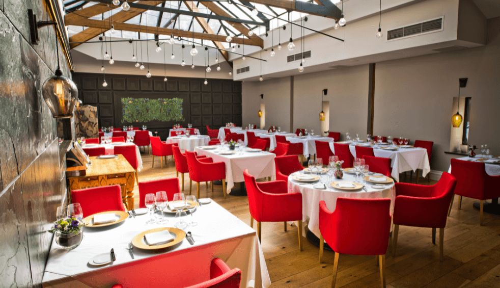 A room in the art school restaurant with lots of tables with white table cloths on and red chairs around them.