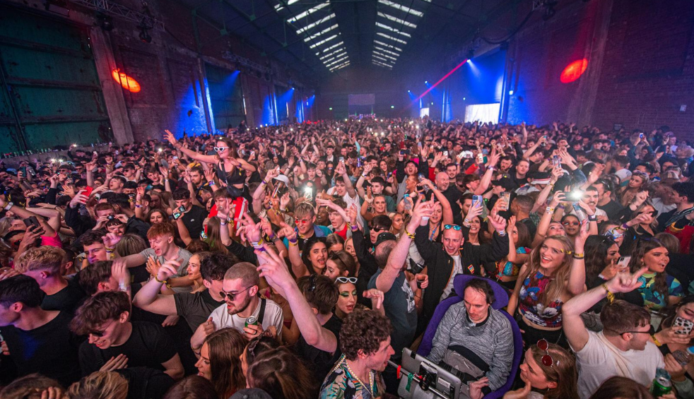 A huge warehouse with neon lights and a huge crowd of people enjoying a gig