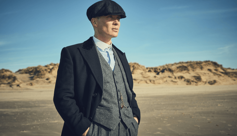 Tommy Shelby wears a suit and flat cap standing on a beach in the TV show Peaky Blinders.