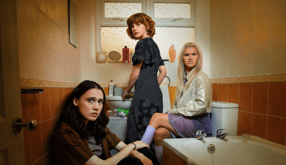 Three girls in a bathroom, two are sitting down and one is standing at the sink