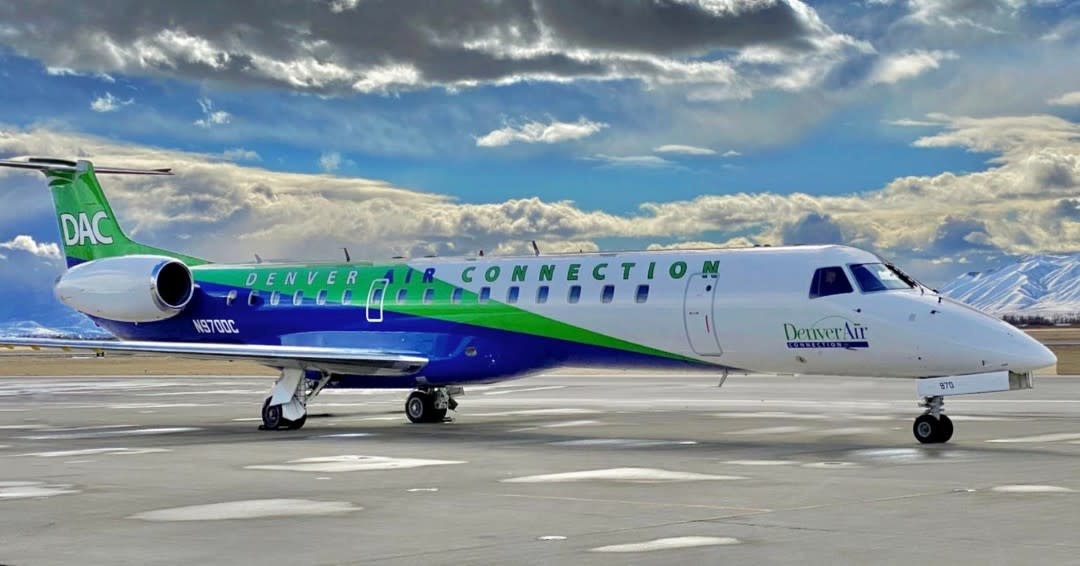 Denver Air Connection aircraft parked on a tarmac, at Muskegon County airport with dramatic mountainous backdrop and partly cloudy sky.