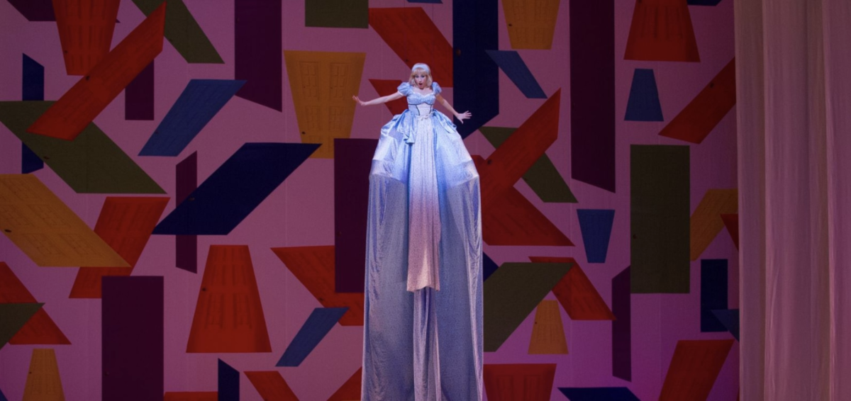 A performer in a light blue ballgown stands elevated against a colorful geometric backdrop, arms outstretched in a theatrical pose. The dramatic costume features an extremely long flowing train extending downward, creating a striking visual contrast with the angular pink, red, blue, and orange shapes behind.