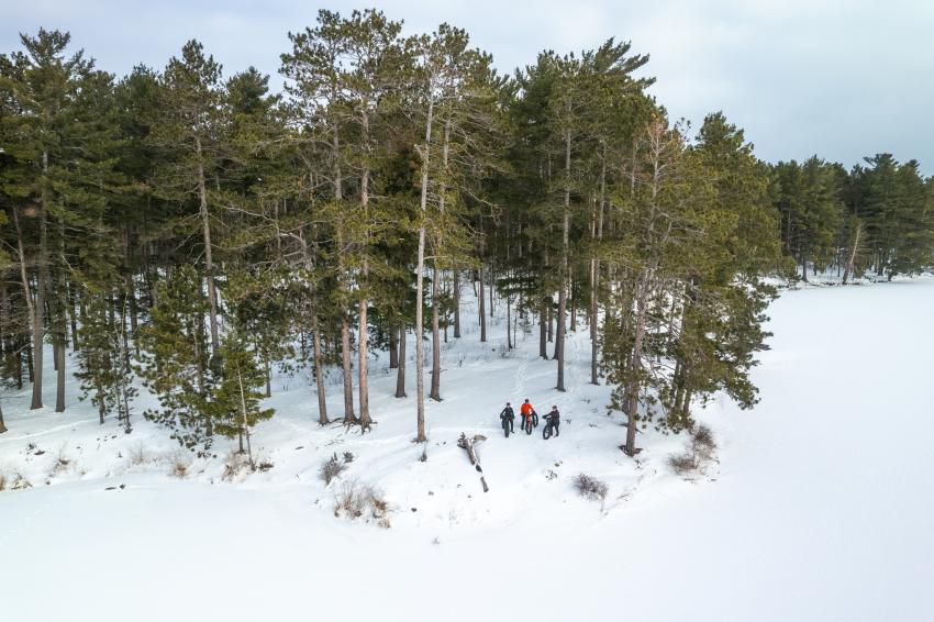 An aerial view of a snowy landscape and evergreen trees featuring three bikers on the NTN trails in Marquette, MI