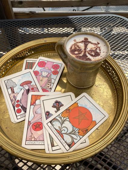 A latte with a set of scales designed into the foam sits on a gold tray with several tarot cards around it.