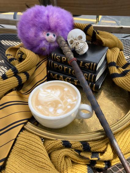 A latte in a white mug sits on a silver tray with a stack of three black books. A yellow and black scrarf and tie surround the tray, a wand lays across it, and a purple stuffed animal sits at the top of the frame. A plastic skull sits on the books.