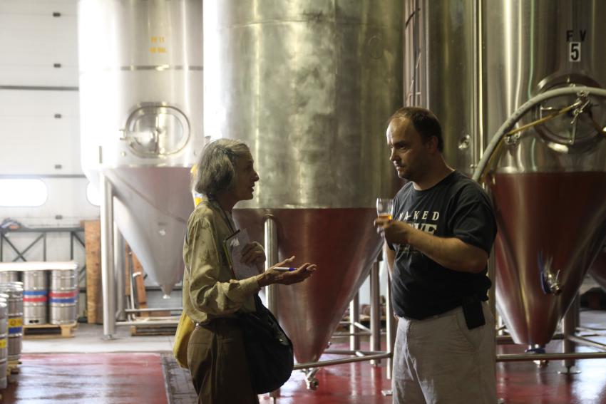 A traveler speaks with the brewmaster at Naked Dove Brewing with large, steel brewing equipment in the background.