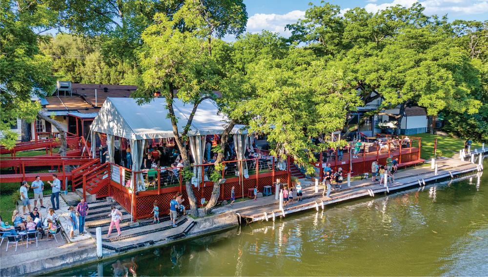 The outdoor lakeside deck at County Line on The Lake with people walking along the deck and large green trees leaning over the deck and seating area.