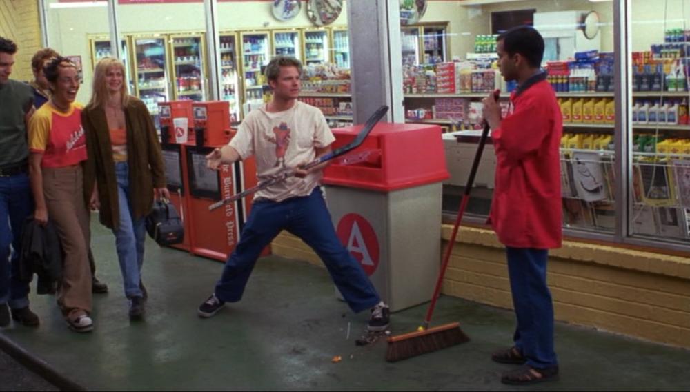 Suburbia screengrab, showing a man in a red shirt sweeping the ground while another man stands in front of him with a hockey stick in his hand. A group of people are walking past them and laughing