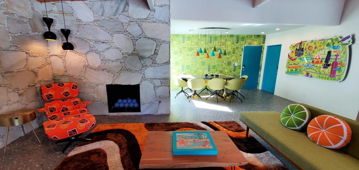 A colorful mid-century modern living room featuring a stone fireplace with blue balls, an orange patterned lounge chair, a green bench with citrus slice pillows, and a round wooden coffee table. There's a dining area with yellow chairs in the background against a vibrant wallpaper with abstract designs. Blue doors and eclectic lighting fixtures complete the retro ambiance.
