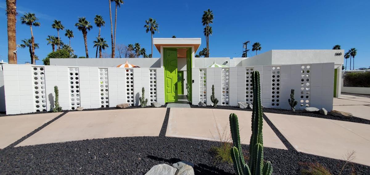 A mid-century modern home exterior with a bright lime green door, decorative breeze blocks, and cactus landscaping, set against a backdrop of tall palm trees under a clear blue sky.