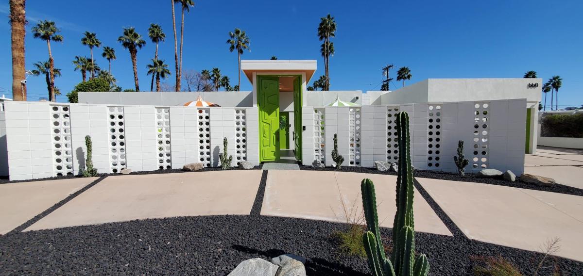A mid-century modern home exterior with a bright lime green door, decorative breeze blocks, and cactus landscaping, set against a backdrop of tall palm trees under a clear blue sky.