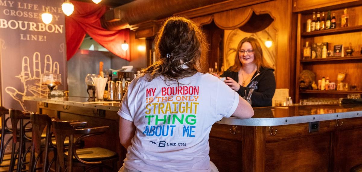 A woman wearing a t-shirt that reads in rainbow letters "My bourbon is the only straight thing about me" seated at the bar at Second Sight Spirits with a woman bartender offering her a drink.