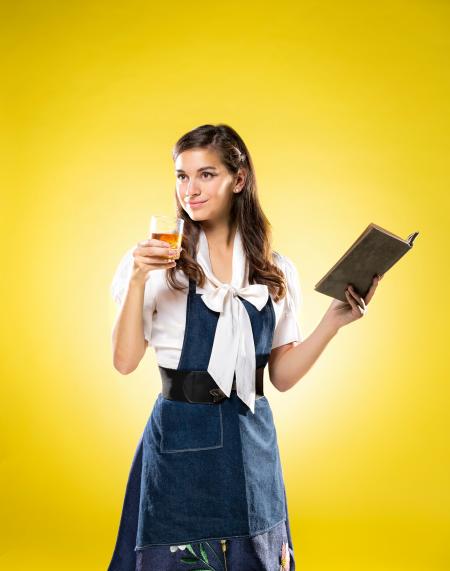 yellow background and woman holding a book in one hand and a bourbon glass in the other.  promotional for beauty and the bourbon tasting event.