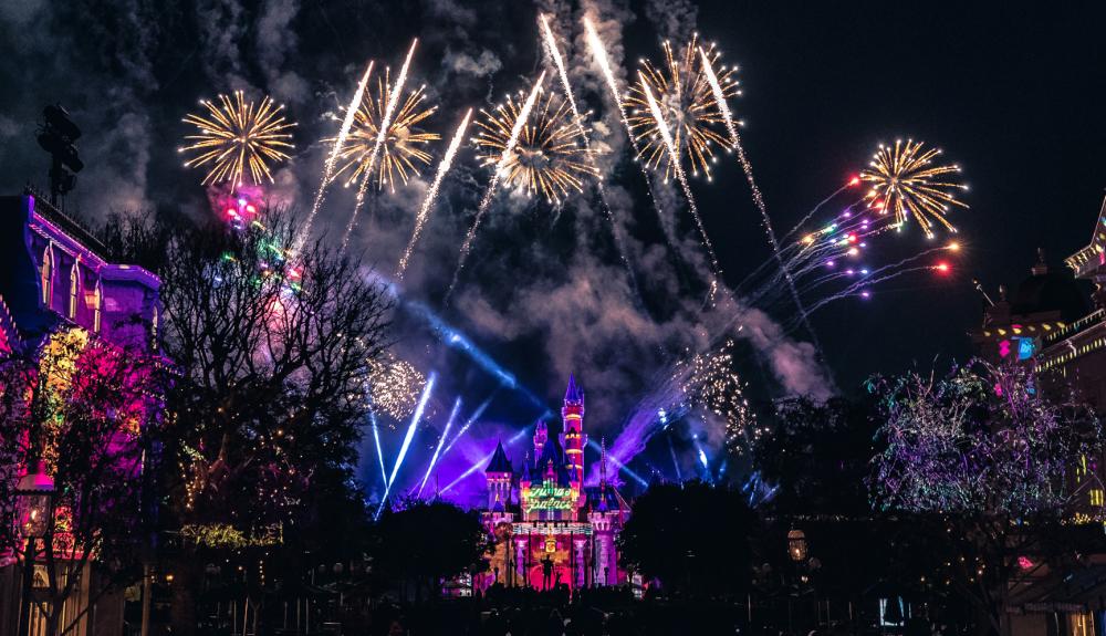 Image of the Sleeping Beauty Castle at Disneyland. Yellow and sparkly fireworks can in the sky behind the castle. Various purple, blue, and white lights can be seen on the castle.